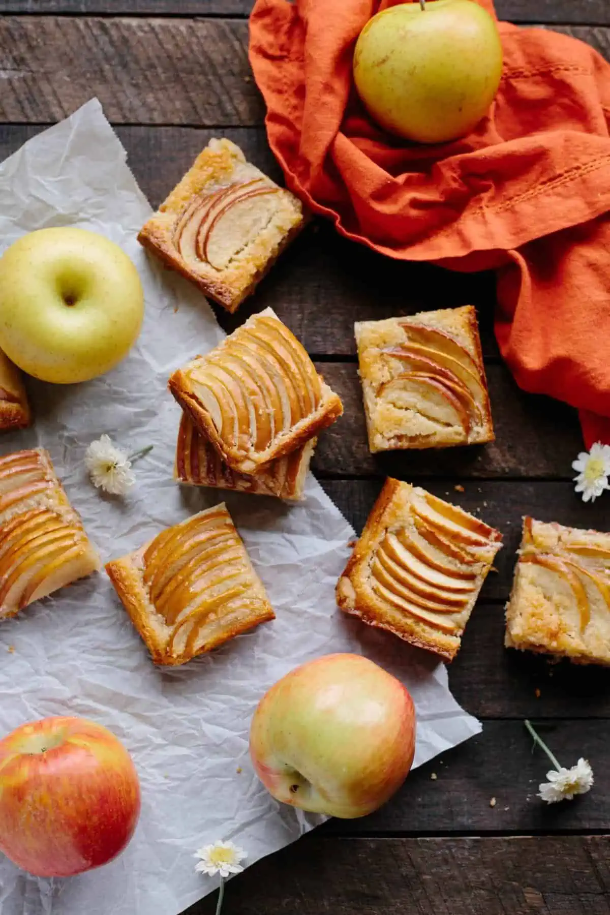 Square slices of almond apple shortbread spread on a table with whole apples mixed in.
