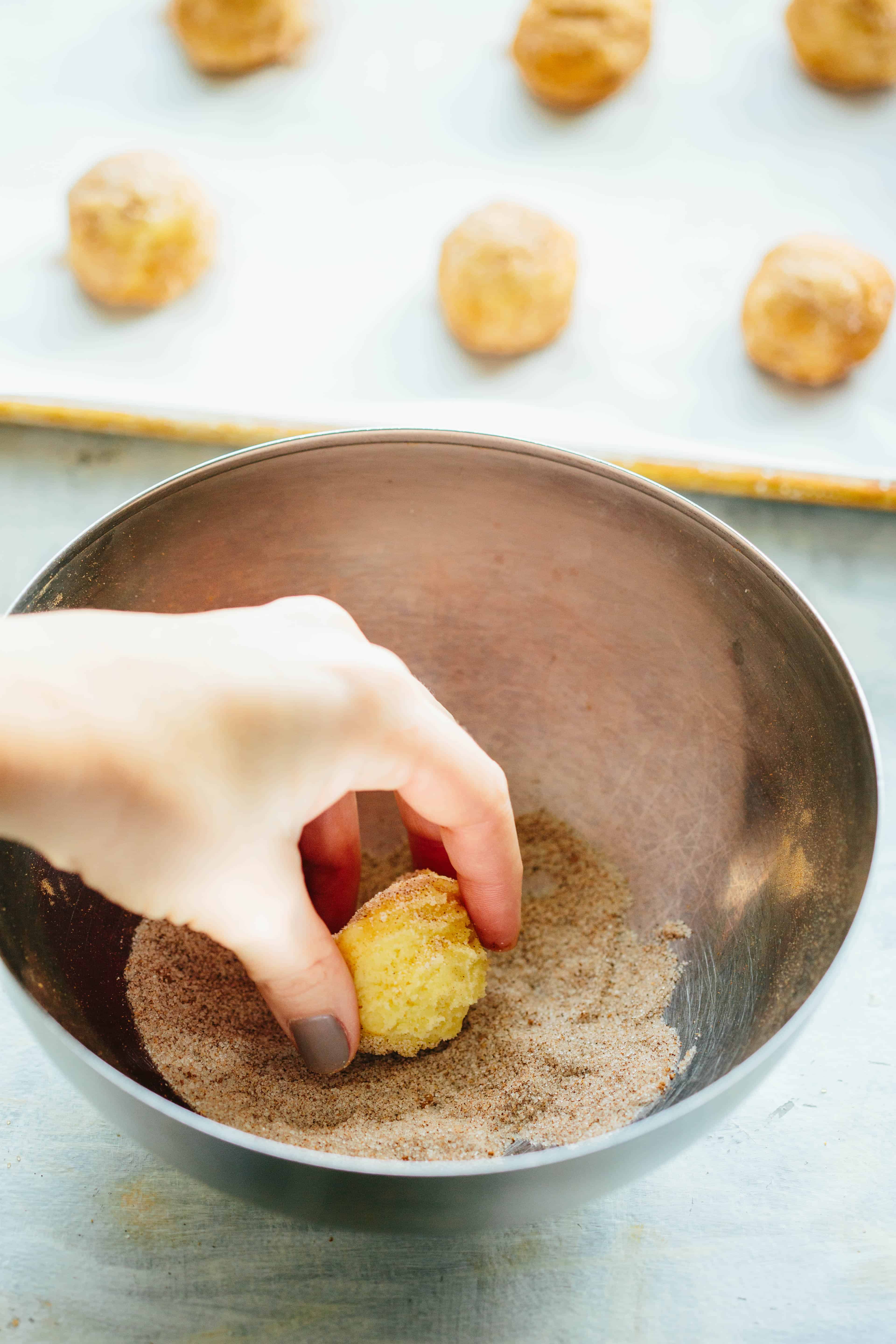 A hand rolling a ball of snickerdoodle dough in a bowl of cinnamon sugar.