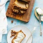 Loaf of Paleo banana bread sliced on a wood cutting board, next to a plate containing a buttered slice.