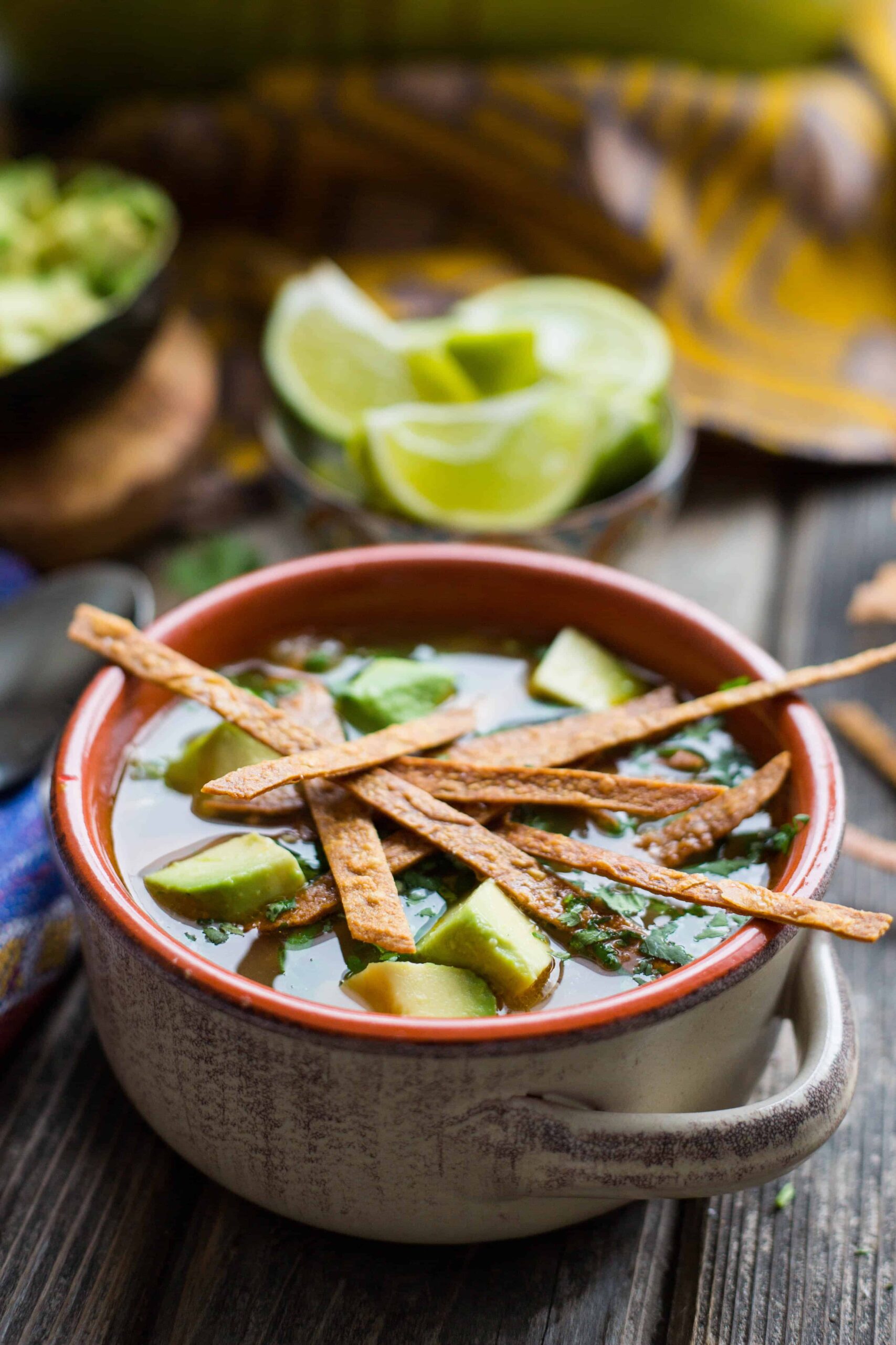 A brown bowl of chicken tortilla soup topped with diced avocado and fried tortilla strips.
