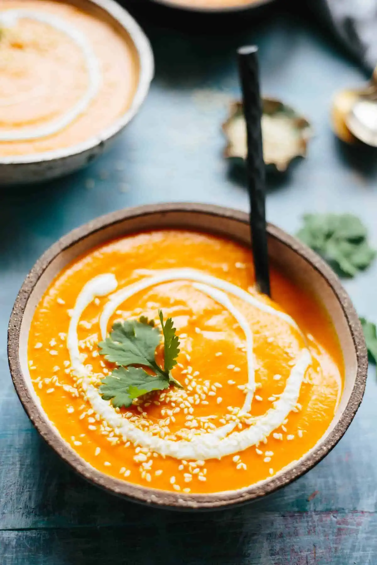 Vertical shot of a bowl of spicy pureed carrot soup garnished with cilantro, sesame seeds, and a yogurt swirl.