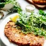 Super close up shot of a breaded chicken cutlet and arugula salad on a white dinner plate.