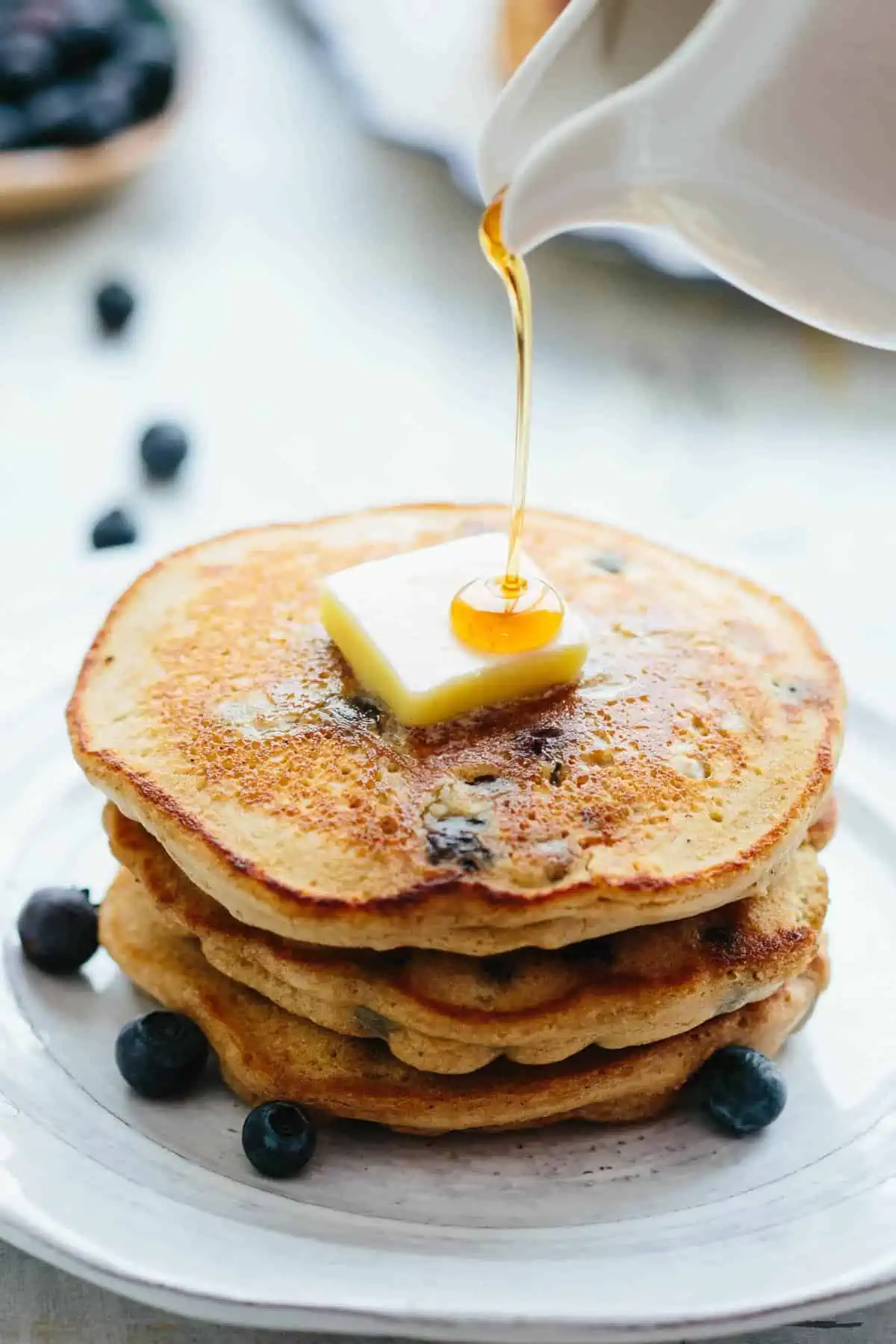 Close up of syrup drizzling onto a butter pat atop a stack of blueberry oatmeal pancakes.