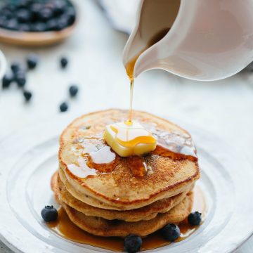 Pouring maple syrup over a stack of blueberry oatmeal pancakes on a plate.