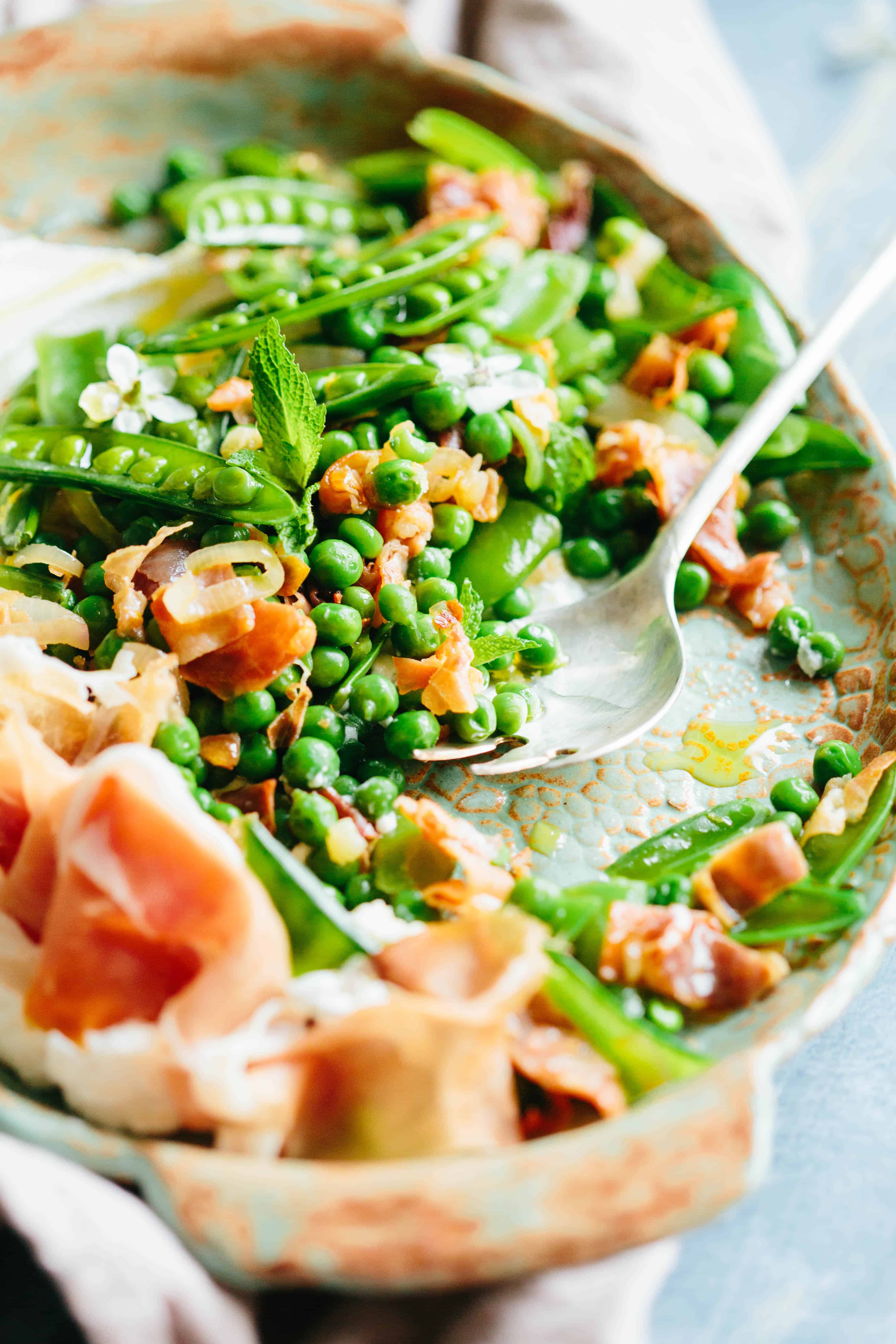 Close up of a serving platter of fresh peas in their pods alongside ribbons of prosciutto.