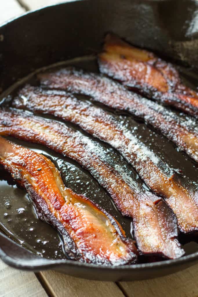 Close up of bacon slices frying in a cast iron skillet.