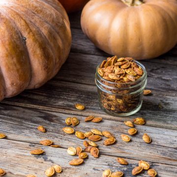 small jar of roasted pumpkin seeds on wood table with seeds scattered around and tan cinderella pumpkins in the background