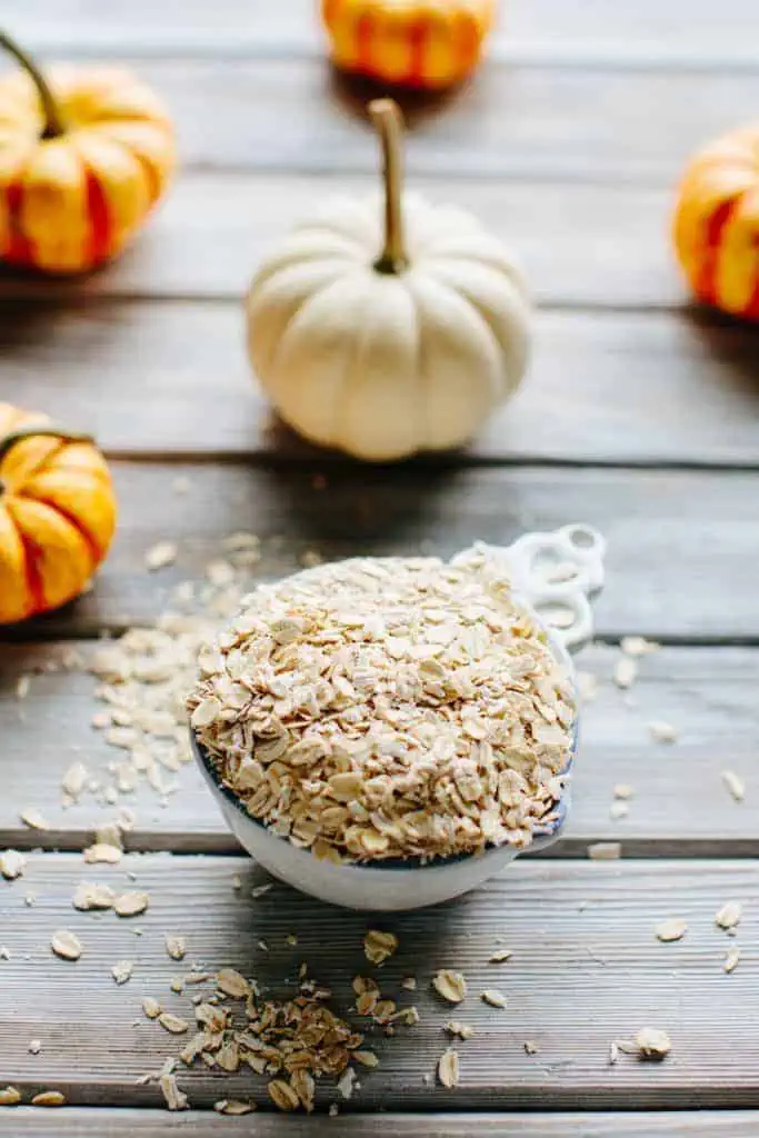 A ceramic measuring cup overflowing with oats on a wooden background with mini pumpkins