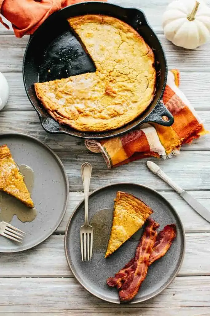 two dark plates with slices of pumpkin dutch baby plus a skillet with remaining pumpkin dutch baby and an orange plaid napkin