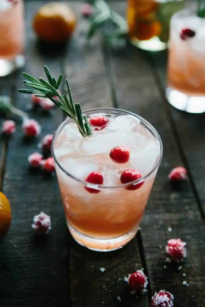 glass of light pink drink with ice, fresh cranberries and rosemary on dark wooden background 