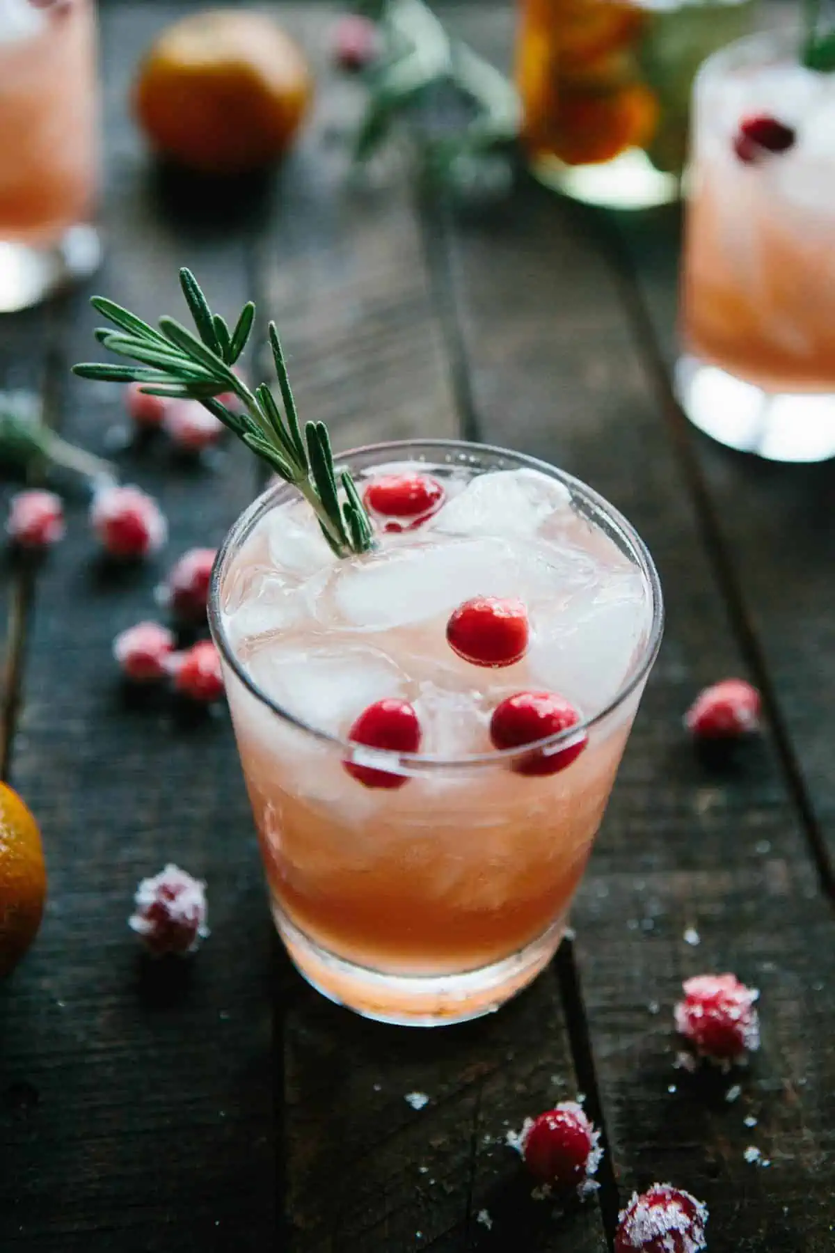 glass of light pink drink with ice, fresh cranberries and rosemary on dark wooden background
