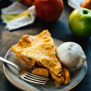 slice of apple pie on a gray plate with a fork and scoop of ice cream with apples in the background