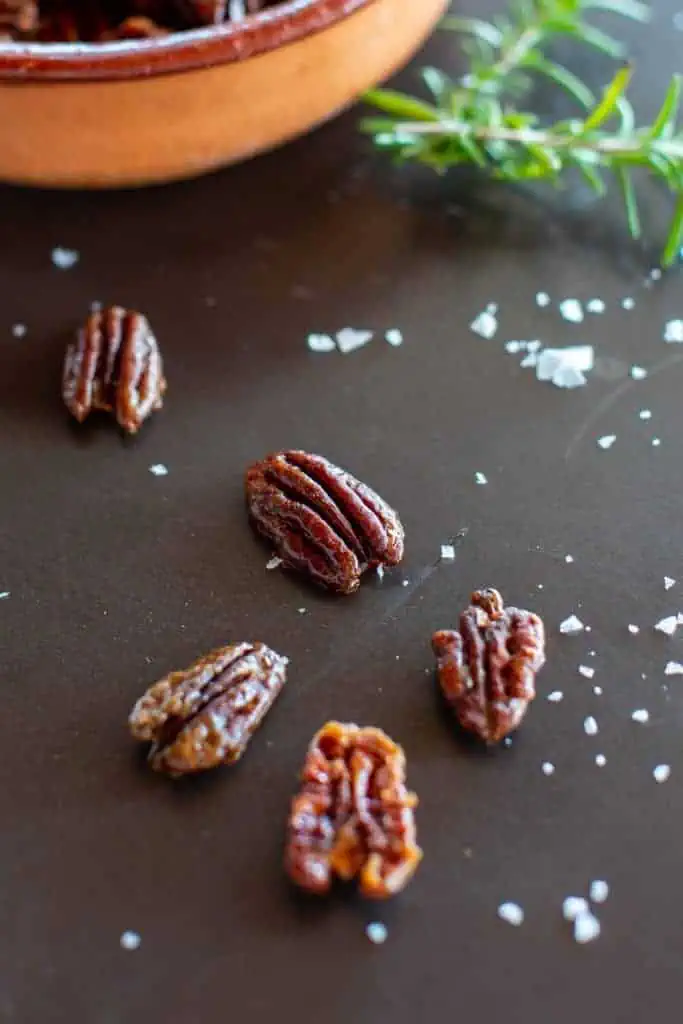 close up of a few rosemary maple roasted pecans on a dark background with sea salt