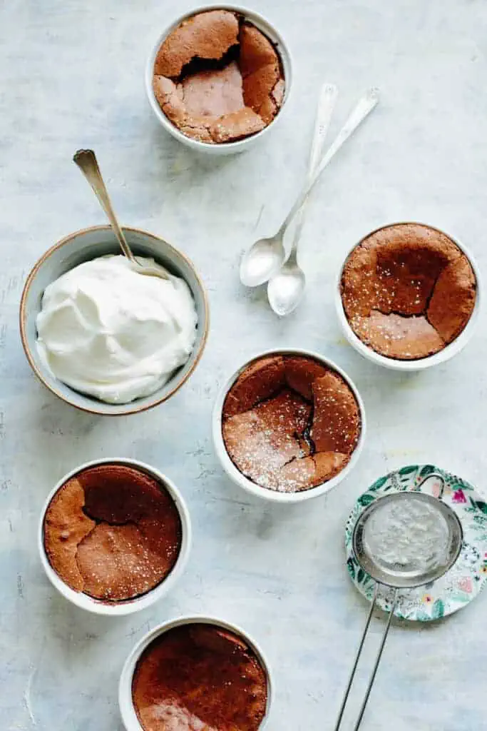 Three mini chocolate souffles in ramekins next to a bowl of freshly whipped cream.