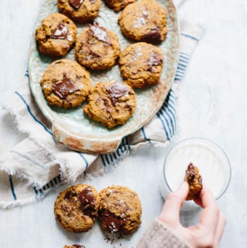 A hand dunking a chocolate chip cookie into a glass of milk next to a serving platter of more cookies.