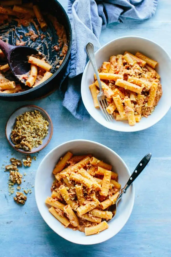 an overhead shot of a table with 2 bowls and a pan of rigatoni, some chopped walnuts, forks and a napkin