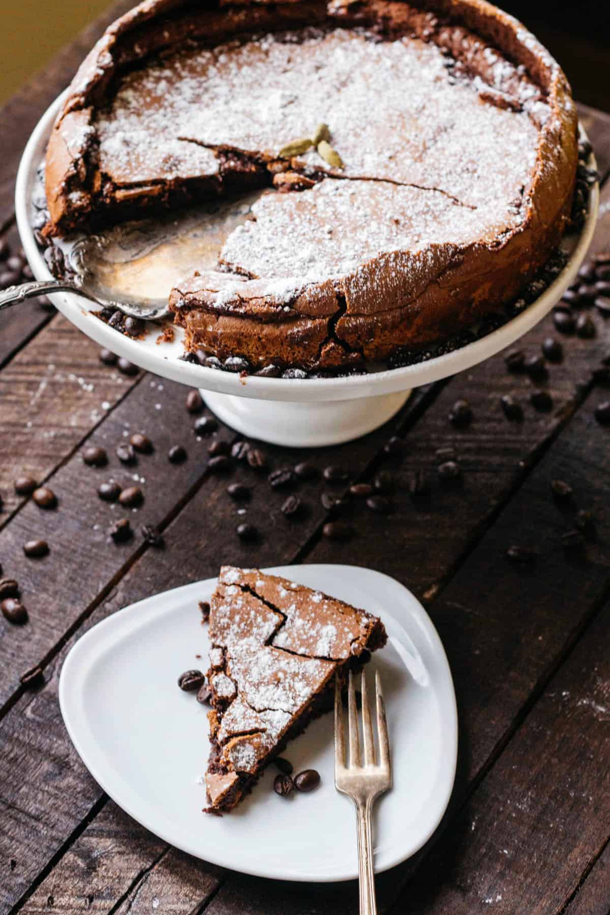 A cake stand holding a coffee flourless chocolate cake next to a dessert plate holding a slice