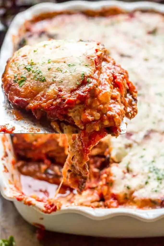 A spatula with a slice of eggplant parmesan with cheese stringing down to a baking dish.
