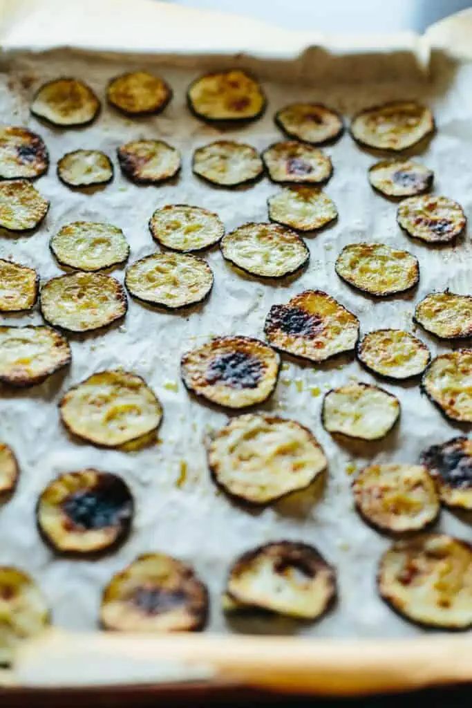 Close up of thin slices of zucchini roasted on a parchment lined baking sheet.