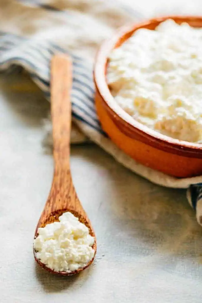 A red bowl filled with homemade ricotta cheese and a wood spoon on the table with more cheese.