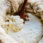 A wood spoon scooping fresh ricotta draining in cheesecloth.