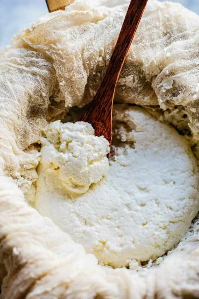 A wood spoon scooping fresh ricotta draining in cheesecloth.