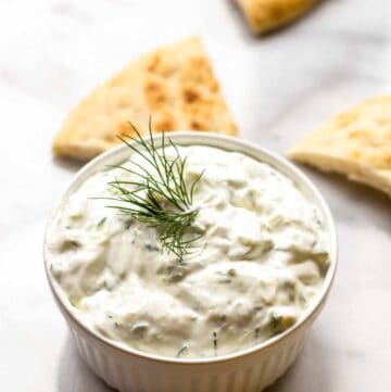 A white ramekin of Tzatziki Sauce with pita triangles in the background.