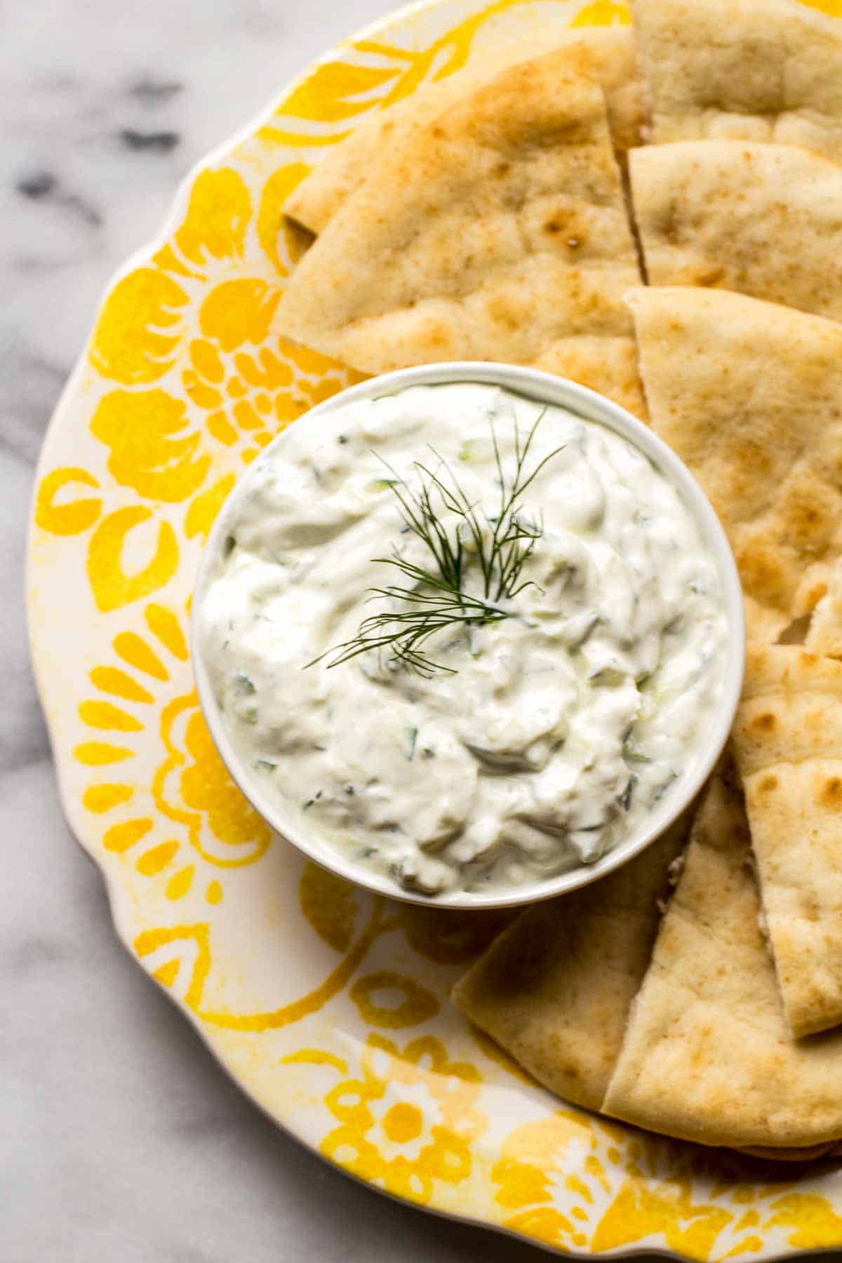Overhead shot of a yellow plate with a bowl of Greek yogurt, dill and cucumber dip and pita wedges.