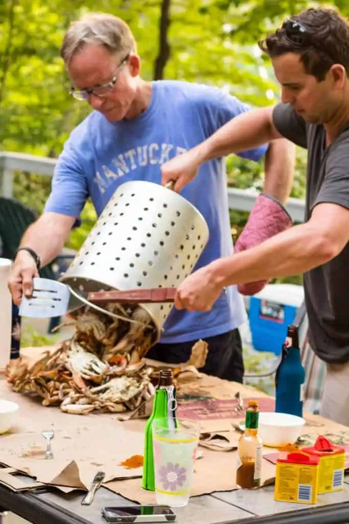 Two men dumping a steamer pot of steamed blue crabs on to a table.
