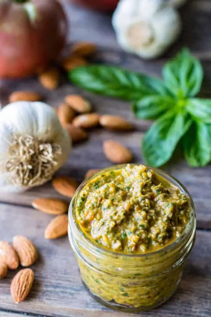 a small jar of pesto on a wood board with whole almonds, garlic and basil in the background. 