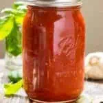 Side view of a large jar of marinara sauce with fresh basil and a head of garlic surrounding it.