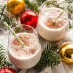 Two glasses of Coquito with cinnamon on top on a bed of Christmas tree branches and red and gold ornaments.