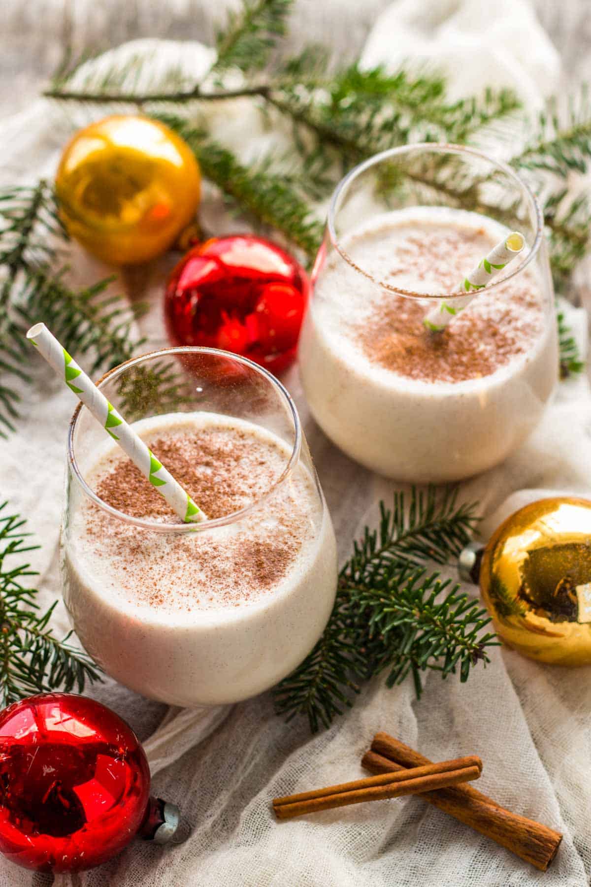 Two glasses of Coquito with cinnamon on top on a bed of Christmas tree branches and red and gold ornaments.