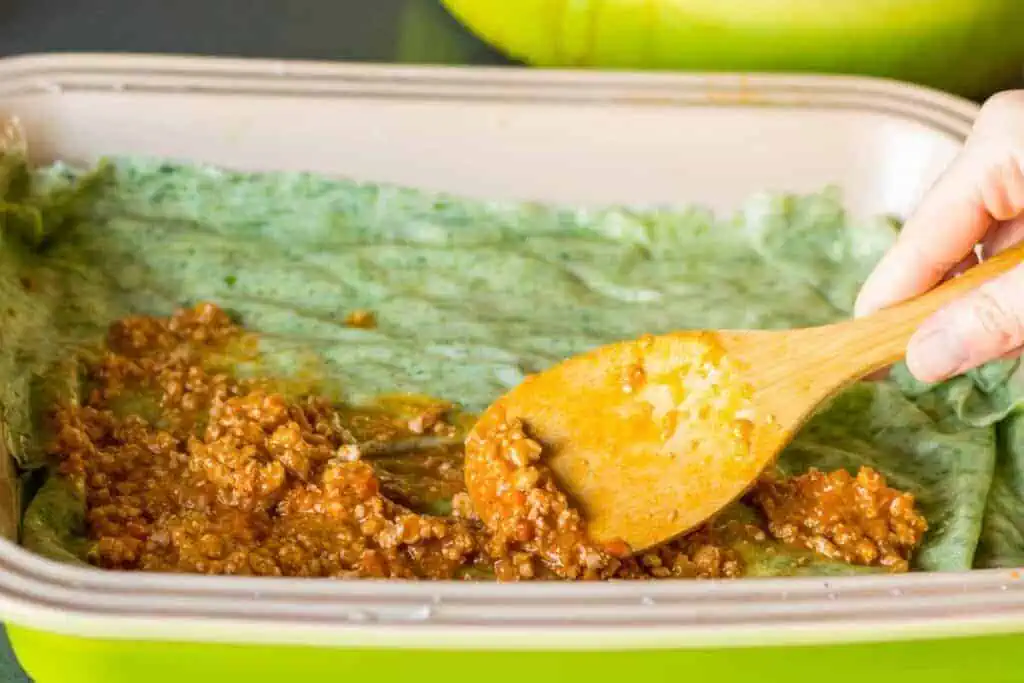 A wooden spoon spreading meat sauce over fresh lasagna noodles in a baking dish.