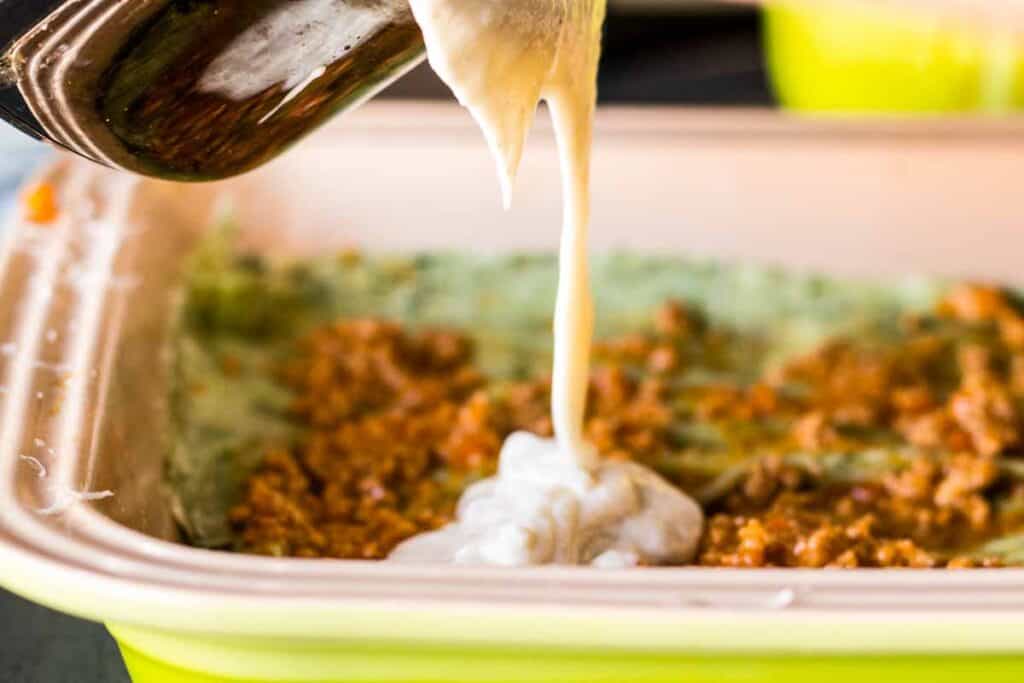 Pouring bechemel sauce from a pot over meat sauce and lasagna noodles in a baking dish.