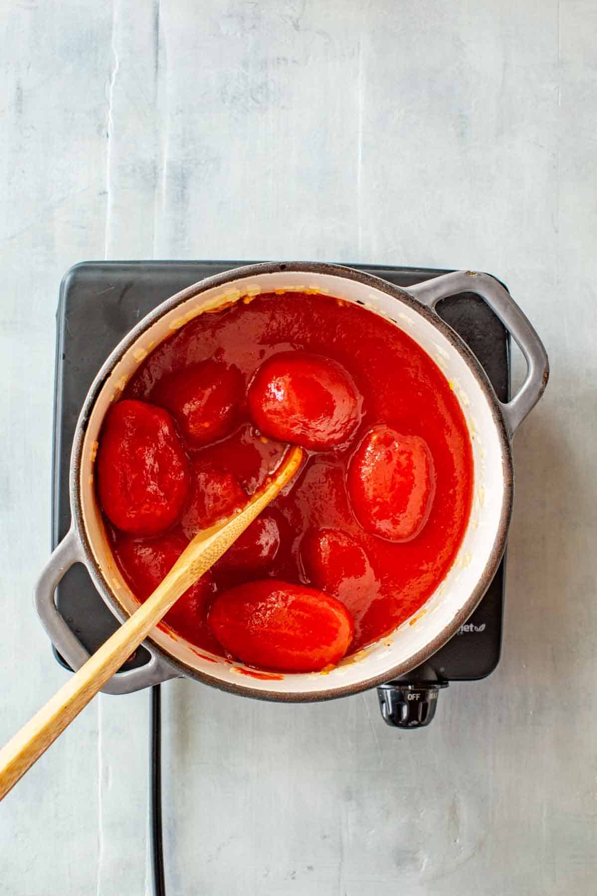 A wooden spoon breaking up canned tomatoes in a pot.