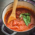 A pot of homemade marinara sauce with a wooden spoon and sprig of basil.