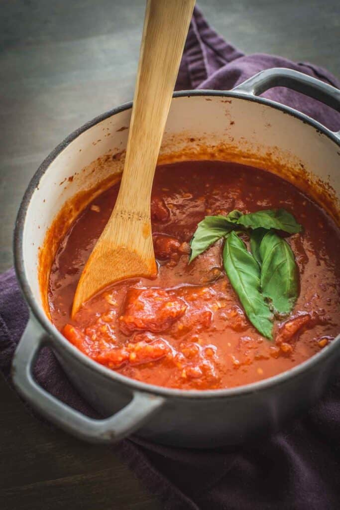 A pot of homemade marinara sauce with a wooden spoon and sprig of basil.