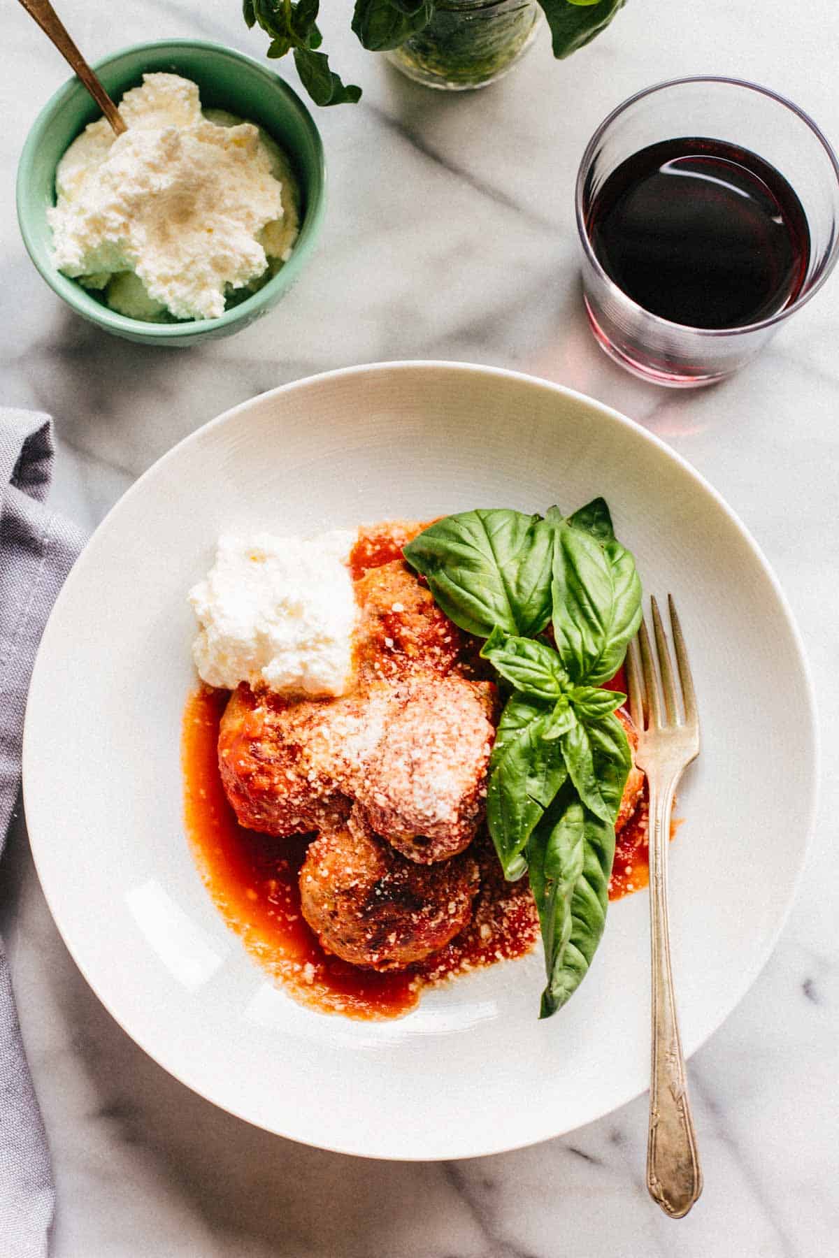 Overhead shot of a white plate with meatballs, a scoop of ricotta, fresh basil and a glass of red wine.