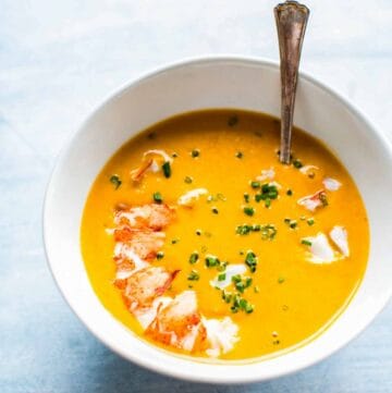 A white bowl with bright orange lobster soup with a lobster tail and chives on top.