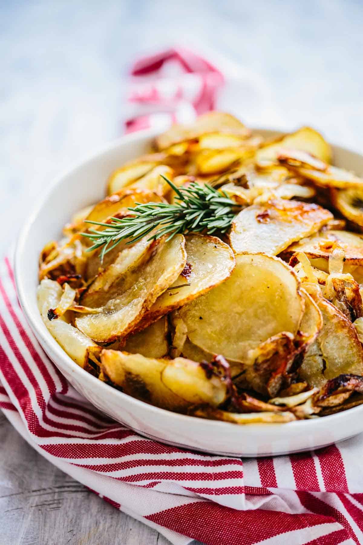 A white bowl of roasted potatoes with a sprig of rosemary on top and a red napkin. 