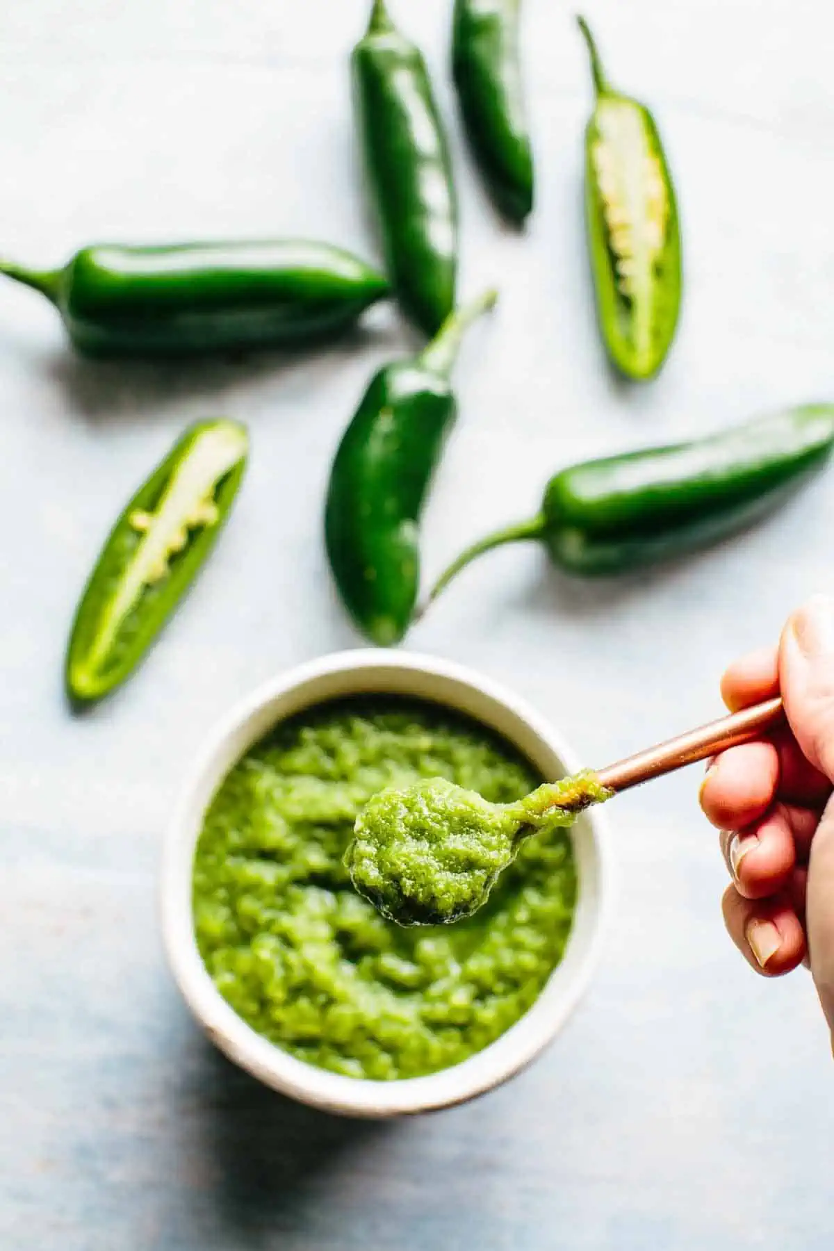 A small bowl of green hot sauce with a hand holding a spoon and jalapeños in the background.