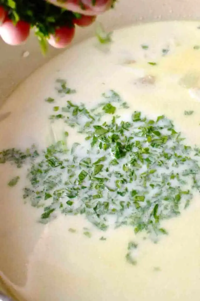 A hand sprinkling parsley into a pot of creamy white soup.