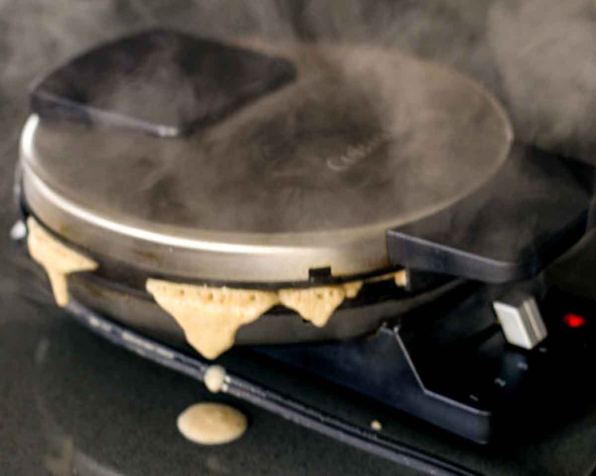 A closed waffle iron with steam and waffle batter coming out.