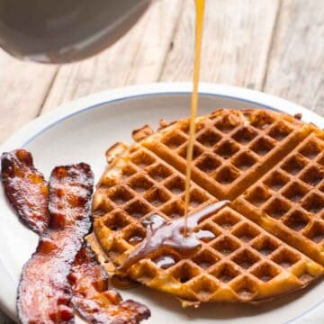 A plate with a waffle and bacon with maple syrup pouring out of a white cup.