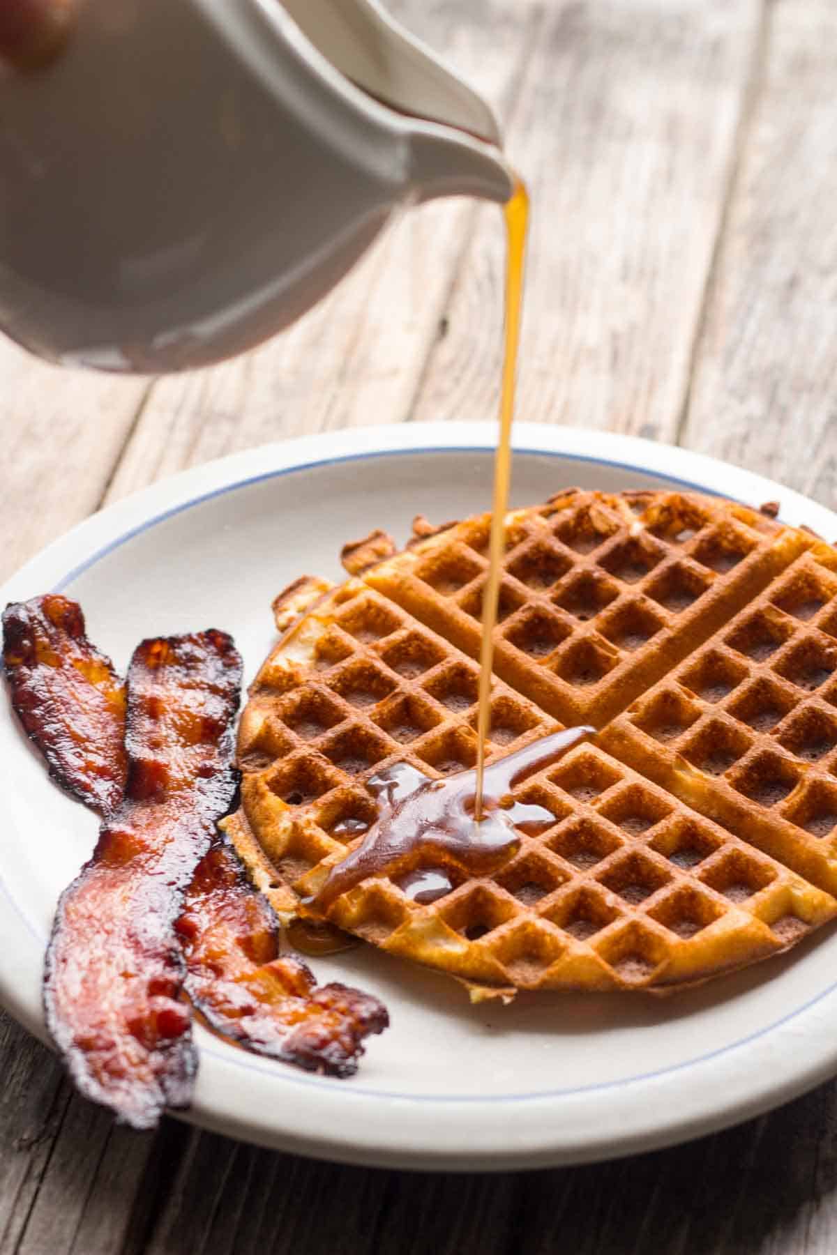 A plate with a waffle and bacon with maple syrup pouring out of a white cup.