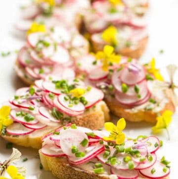 A white platter of toasts topped with thinly sliced radishes, herbs and edible flowers.