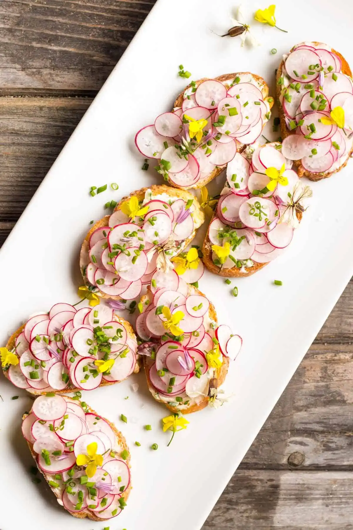 Overhead shot of a white platter of buttered radish toasts topped with fresh  herbs and edible flowers on top.