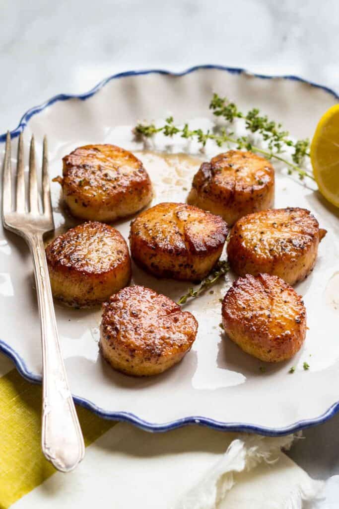 A white plate with pan seared scallops with fresh thyme and a lemon in the background.