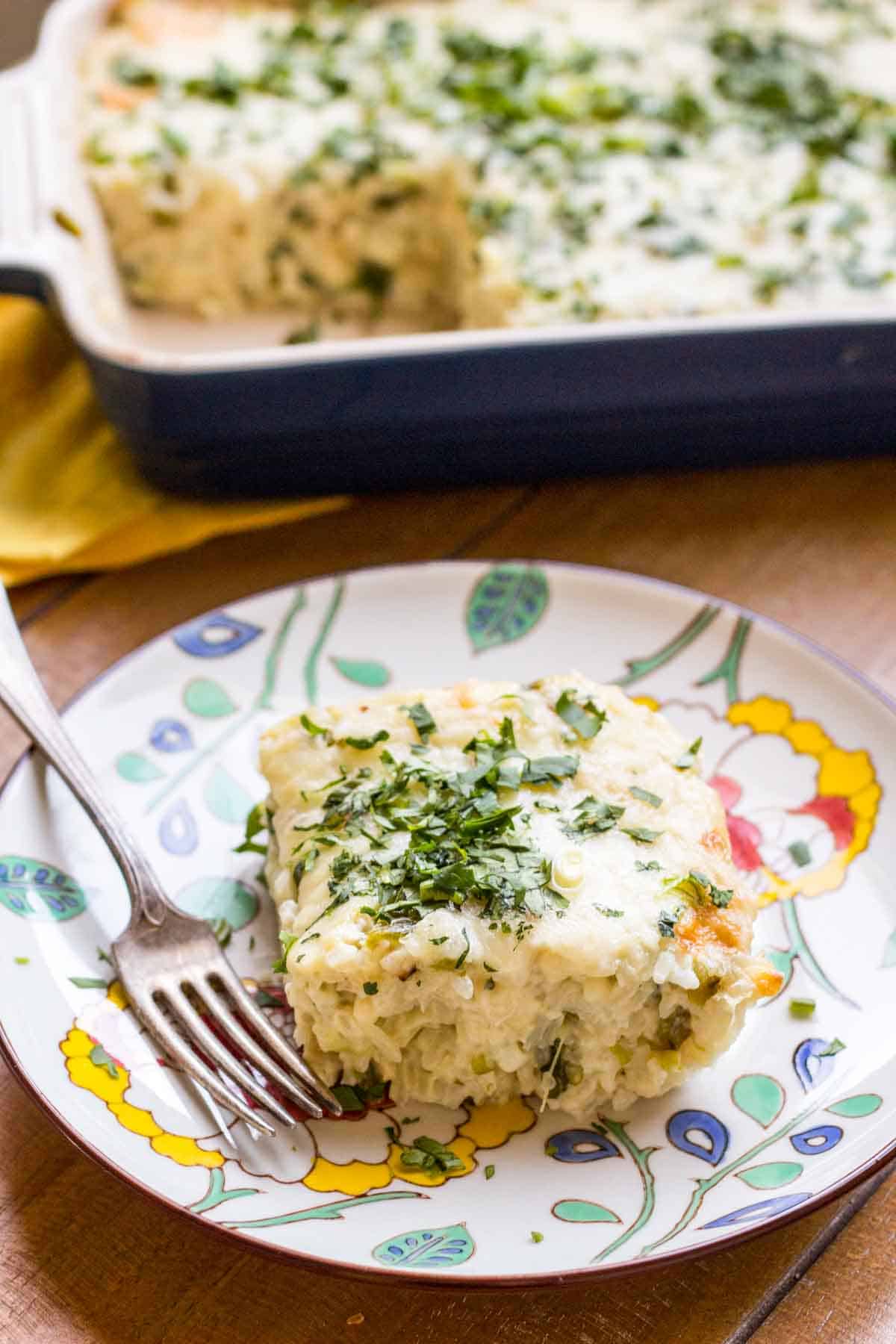 A bright floral plate with a slice of cheesy rice casserole with the dish in the background.